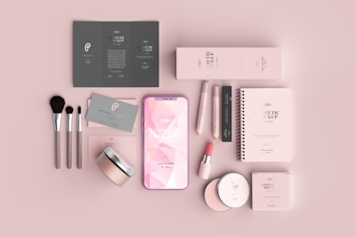 A neatly arranged flat lay of cosmetic products and accessories, including brushes, a smartphone, packaging boxes, and a spiral-bound notebook, all in a soft pink color scheme.