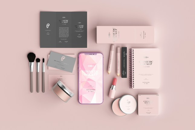 A neatly arranged flat lay of cosmetic products and accessories, including brushes, a smartphone, packaging boxes, and a spiral-bound notebook, all in a soft pink color scheme.