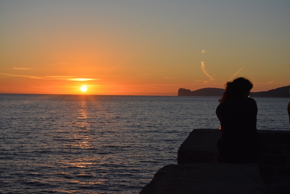 silhouette of woman sitting on rock near body of water during sunset