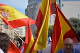 Brightly colored Spanish flags are prominently displayed, with people gathered around in a public space. The setting appears to be a lively event, with flags waving in the air. The background features urban architecture, including a cylindrical building and greenery.