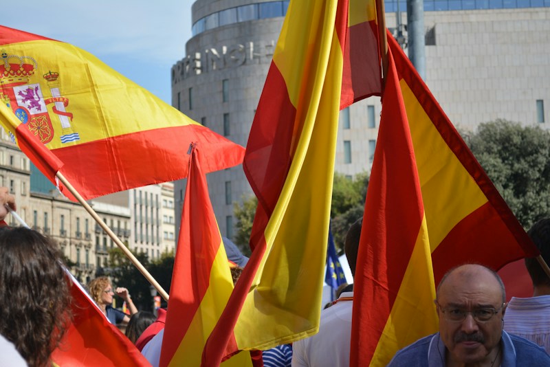 Brightly colored Spanish flags are prominently displayed, with people gathered around in a public space. The setting appears to be a lively event, with flags waving in the air. The background features urban architecture, including a cylindrical building and greenery.