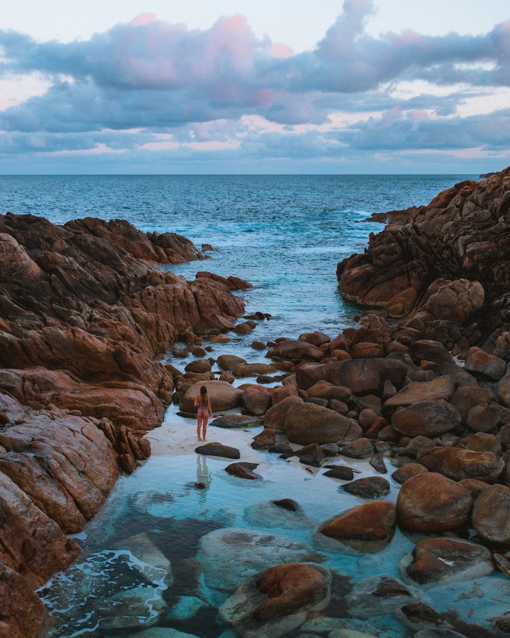 woman in white shirt and blue denim jeans standing on rocky shore during daytime