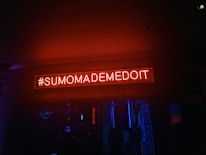 A neon sign with the text '#SUMOMADEMEDOIT' illuminated in red against a dark background. The environment seems like a nightclub or bar setting with dim lighting and blue accents.
