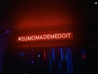 A neon sign with the text '#SUMOMADEMEDOIT' illuminated in red against a dark background. The environment seems like a nightclub or bar setting with dim lighting and blue accents.