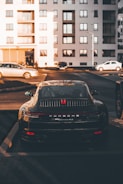 A luxury sports car is parked in a residential area, surrounded by modern apartment buildings. The sleek vehicle is prominently displayed, catching the warm glow of the setting sun. Nearby, other cars are parked, hinting at an urban environment.