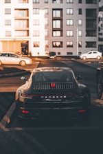 A luxury sports car is parked in a residential area, surrounded by modern apartment buildings. The sleek vehicle is prominently displayed, catching the warm glow of the setting sun. Nearby, other cars are parked, hinting at an urban environment.