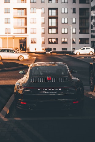 A luxury sports car is parked in a residential area, surrounded by modern apartment buildings. The sleek vehicle is prominently displayed, catching the warm glow of the setting sun. Nearby, other cars are parked, hinting at an urban environment.