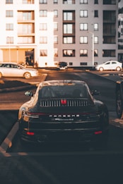 A luxury sports car is parked in a residential area, surrounded by modern apartment buildings. The sleek vehicle is prominently displayed, catching the warm glow of the setting sun. Nearby, other cars are parked, hinting at an urban environment.