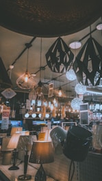 A variety of decorative ceiling and table lamps are displayed in a showroom, with intricate designs and warm lighting. The ceiling features numerous hanging pendant lights in different geometric shapes, and the tables are lined with various lamp models. Price tags and promotional signs are visible throughout the store.