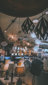 A variety of decorative ceiling and table lamps are displayed in a showroom, with intricate designs and warm lighting. The ceiling features numerous hanging pendant lights in different geometric shapes, and the tables are lined with various lamp models. Price tags and promotional signs are visible throughout the store.