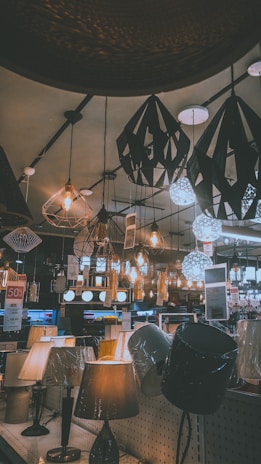 A variety of decorative ceiling and table lamps are displayed in a showroom, with intricate designs and warm lighting. The ceiling features numerous hanging pendant lights in different geometric shapes, and the tables are lined with various lamp models. Price tags and promotional signs are visible throughout the store.