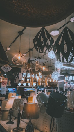 A variety of decorative ceiling and table lamps are displayed in a showroom, with intricate designs and warm lighting. The ceiling features numerous hanging pendant lights in different geometric shapes, and the tables are lined with various lamp models. Price tags and promotional signs are visible throughout the store.