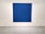 A large, textured blue canvas is mounted on a white gallery wall. Small, evenly spaced perforations create a subtle pattern across the surface. The artwork is cordoned off by a simple barrier with thin ropes attached to white circular stands on the floor.