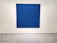 A large, textured blue canvas is mounted on a white gallery wall. Small, evenly spaced perforations create a subtle pattern across the surface. The artwork is cordoned off by a simple barrier with thin ropes attached to white circular stands on the floor.