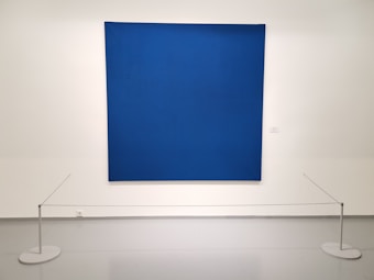A large, textured blue canvas is mounted on a white gallery wall. Small, evenly spaced perforations create a subtle pattern across the surface. The artwork is cordoned off by a simple barrier with thin ropes attached to white circular stands on the floor.