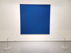 A large, textured blue canvas is mounted on a white gallery wall. Small, evenly spaced perforations create a subtle pattern across the surface. The artwork is cordoned off by a simple barrier with thin ropes attached to white circular stands on the floor.