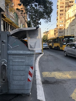 A street scene with a large gray trash bin on the left side of the image, partially open, situated on a sidewalk. In the background, there is a yellow city bus in motion and a parked gray car. The street is lined with tall buildings, some with scaffolding. Trees and overhead wires are visible, suggesting an urban environment.