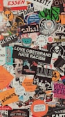 A collage of various colorful stickers layered over each other. The stickers have diverse themes including music, activism, brands, and art. Some include phrases like 'Love Obstbrand Hate Racism' and 'Feminist Pursuits'. The composition is vibrant and chaotic, featuring a mix of typography and graphic designs.