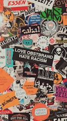 A collage of various colorful stickers layered over each other. The stickers have diverse themes including music, activism, brands, and art. Some include phrases like 'Love Obstbrand Hate Racism' and 'Feminist Pursuits'. The composition is vibrant and chaotic, featuring a mix of typography and graphic designs.