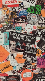 A collage of various colorful stickers layered over each other. The stickers have diverse themes including music, activism, brands, and art. Some include phrases like 'Love Obstbrand Hate Racism' and 'Feminist Pursuits'. The composition is vibrant and chaotic, featuring a mix of typography and graphic designs.
