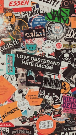 A collage of various colorful stickers layered over each other. The stickers have diverse themes including music, activism, brands, and art. Some include phrases like 'Love Obstbrand Hate Racism' and 'Feminist Pursuits'. The composition is vibrant and chaotic, featuring a mix of typography and graphic designs.