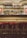 A dimly lit bar with shelves filled with a variety of liquor bottles. The bar features three red cushioned stools and is adorned with warm wall lamps on either side, adding a cozy and vintage feel. The arching stone ceiling adds to the rustic ambiance.
