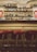 A dimly lit bar with shelves filled with a variety of liquor bottles. The bar features three red cushioned stools and is adorned with warm wall lamps on either side, adding a cozy and vintage feel. The arching stone ceiling adds to the rustic ambiance.