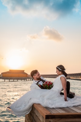 A couple dressed in wedding attire sits on a wooden dock by the water during sunset. The groom wears a tuxedo and holds the bride, who is in a white gown with a lace design. She holds a bouquet of red flowers. The background features calm water and overwater bungalows against a sunset sky.