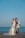 A couple is dressed in wedding attire, with the bride wearing a white gown and the groom in a suit. They are sharing a kiss by the seaside, holding a vintage-style bicycle with a basket of red and white flowers.