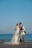 A couple is dressed in wedding attire, with the bride wearing a white gown and the groom in a suit. They are sharing a kiss by the seaside, holding a vintage-style bicycle with a basket of red and white flowers.