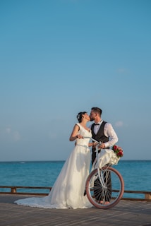 A couple is dressed in wedding attire, with the bride wearing a white gown and the groom in a suit. They are sharing a kiss by the seaside, holding a vintage-style bicycle with a basket of red and white flowers.