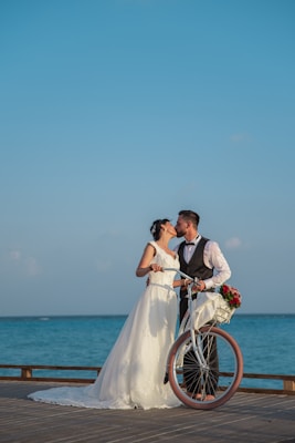 A couple is dressed in wedding attire, with the bride wearing a white gown and the groom in a suit. They are sharing a kiss by the seaside, holding a vintage-style bicycle with a basket of red and white flowers.