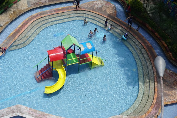 A children's pool with a brightly colored play structure featuring slides in red, blue, green, and yellow. Several children are playing and swimming around the structure, enjoying a sunny day in the water. The pool has a gradual depth with tiered levels and is surrounded by a paved area.
