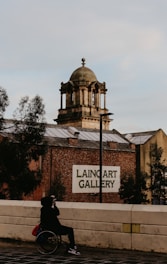 A person in a wheelchair is taking a photo with a smartphone while positioned on a pathway. The background features a historic building with a prominent dome and the sign 'Laing Art Gallery' on the facade. The scene is framed by some trees, and the atmosphere suggests early morning or late afternoon with a subdued lighting.