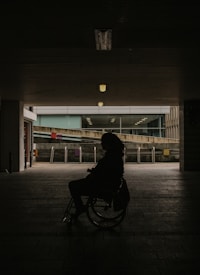 A silhouette of a person in a wheelchair set against a dimly lit tunnel with an urban backdrop visible in the distance. Overhead lights cast a subtle glow, partially illuminating the brick pathway.