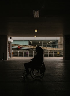 A silhouette of a person in a wheelchair set against a dimly lit tunnel with an urban backdrop visible in the distance. Overhead lights cast a subtle glow, partially illuminating the brick pathway.