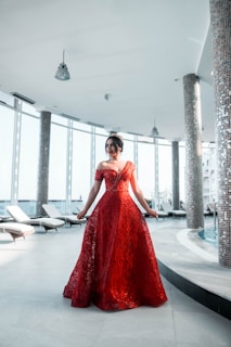 A woman wearing an elegant red gown with an intricate lace design stands in a spacious, modern indoor setting. The room features high ceilings, large windows allowing natural light to fill the space, and several lounge chairs lined up along the windows. The floor is made of light-colored tiles and there are cylindrical pillars covered in small, reflective tiles.