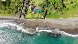Aerial view of a tropical beach scene featuring a vibrant turquoise sea meeting a sandy shore, with waves gently crashing. Above the beach, a lush green area with numerous palm trees accompanies a uniquely shaped swimming pool, part of a landscaped property.