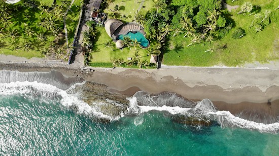 Aerial view of a tropical beach scene featuring a vibrant turquoise sea meeting a sandy shore, with waves gently crashing. Above the beach, a lush green area with numerous palm trees accompanies a uniquely shaped swimming pool, part of a landscaped property.