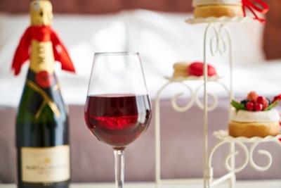 A glass of red wine is prominently placed in the foreground, surrounded by a bottle of champagne with a red ribbon tied around its neck, and an elegant stand holding pastries decorated with fruit and macarons. The setting suggests a sophisticated and indulgent atmosphere, possibly in a bedroom with a neatly made bed in the background.