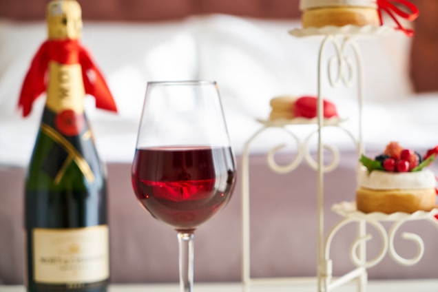 A glass of red wine is prominently placed in the foreground, surrounded by a bottle of champagne with a red ribbon tied around its neck, and an elegant stand holding pastries decorated with fruit and macarons. The setting suggests a sophisticated and indulgent atmosphere, possibly in a bedroom with a neatly made bed in the background.