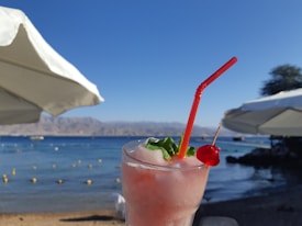 A pink frozen cocktail with a red straw and a cherry garnish sits in the foreground, against a backdrop of a sunny beach scene with azure water and a clear sky. Two white beach umbrellas provide partial shade.