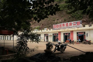 Several motorcycles are parked in front of a building with signs written in Chinese characters. The building appears to be situated in a rural setting, surrounded by trees and a hillside. There is a dirt road or open ground in front of the building.