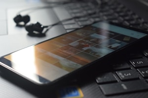 A smartphone displaying a gallery of images rests on a laptop keyboard. The lighting creates reflections on the phone screen. Black earphones are placed beside the smartphone, and the overall scene gives a sense of technology and connectivity.