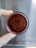 A gloved hand holds a petri dish with a red medium inside, featuring a playful drawing of a smiling face using a dark substance.