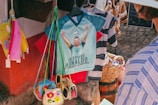 A market stall displays a colorful collection of items including shirts, bags, and accessories. A prominent shirt features a graphic of a soccer player with raised arms and text below, while a wicker basket and various patterned shirts hang nearby. Bright colors such as red, pink, yellow, and blue are visible, with a cobblestone ground and part of a person wearing a striped shirt in the foreground.