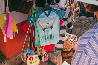 A market stall displays a colorful collection of items including shirts, bags, and accessories. A prominent shirt features a graphic of a soccer player with raised arms and text below, while a wicker basket and various patterned shirts hang nearby. Bright colors such as red, pink, yellow, and blue are visible, with a cobblestone ground and part of a person wearing a striped shirt in the foreground.