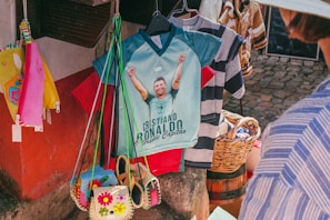 A market stall displays a colorful collection of items including shirts, bags, and accessories. A prominent shirt features a graphic of a soccer player with raised arms and text below, while a wicker basket and various patterned shirts hang nearby. Bright colors such as red, pink, yellow, and blue are visible, with a cobblestone ground and part of a person wearing a striped shirt in the foreground.