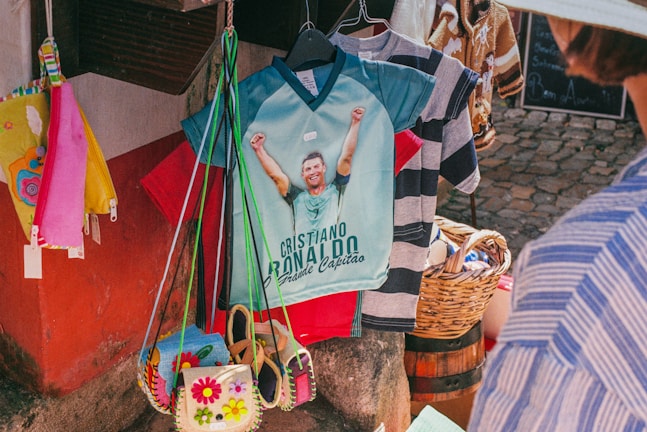 A market stall displays a colorful collection of items including shirts, bags, and accessories. A prominent shirt features a graphic of a soccer player with raised arms and text below, while a wicker basket and various patterned shirts hang nearby. Bright colors such as red, pink, yellow, and blue are visible, with a cobblestone ground and part of a person wearing a striped shirt in the foreground.
