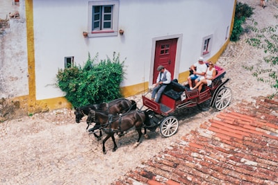 A horse-drawn carriage with a driver and two passengers traverses a cobblestone street adjacent to a rounded corner building with white walls and yellow accents. The carriage is a deep red with white wheels, harnessed to two black horses. Lush green shrubs are visible against the building, and the roof on the right side features red tiles.